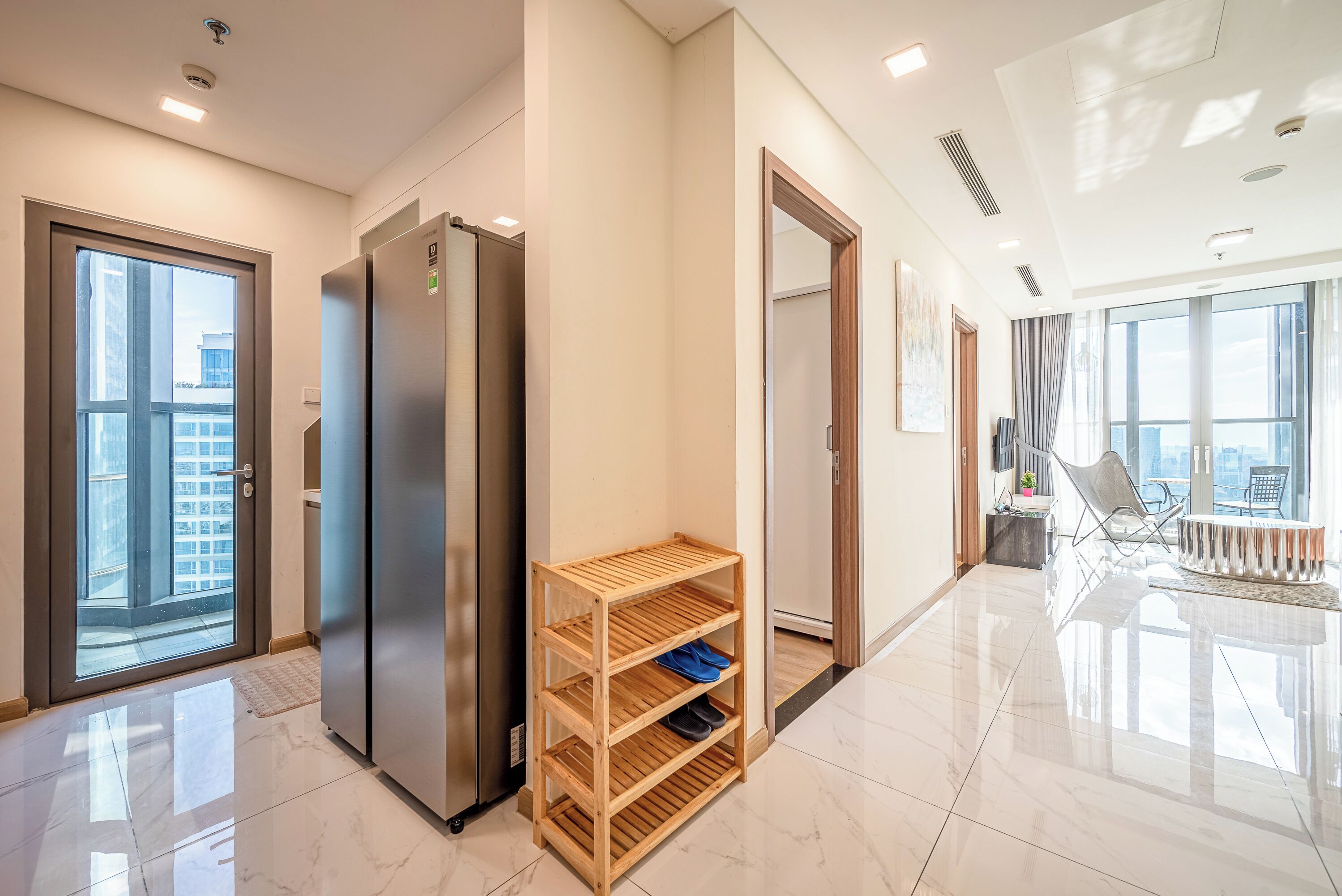 Habitación de hotel moderna con un ventanal grande, refrigerador integrado, estante de madera y zona de estar con vistas a la ciudad.