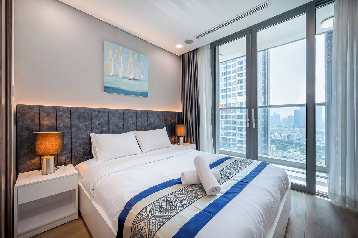 A modern hotel room with a large bed, a painting of sailboats, a city view, and a nightstand with a lamp.