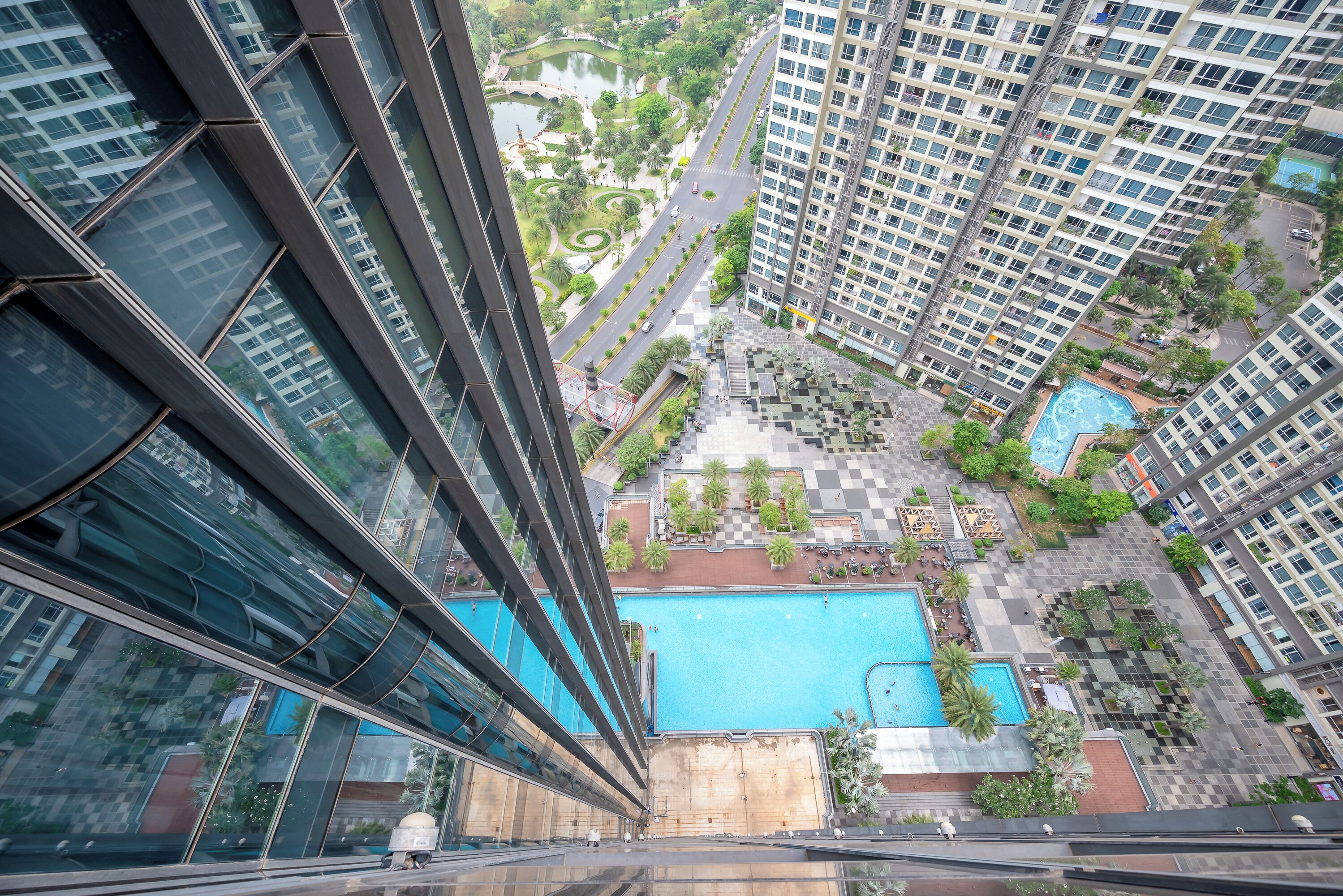 Un immeuble de grande hauteur doté d’un espace piscine comprenant une grande piscine et une plus petite, le tout entouré de verdure et bordé par une route.