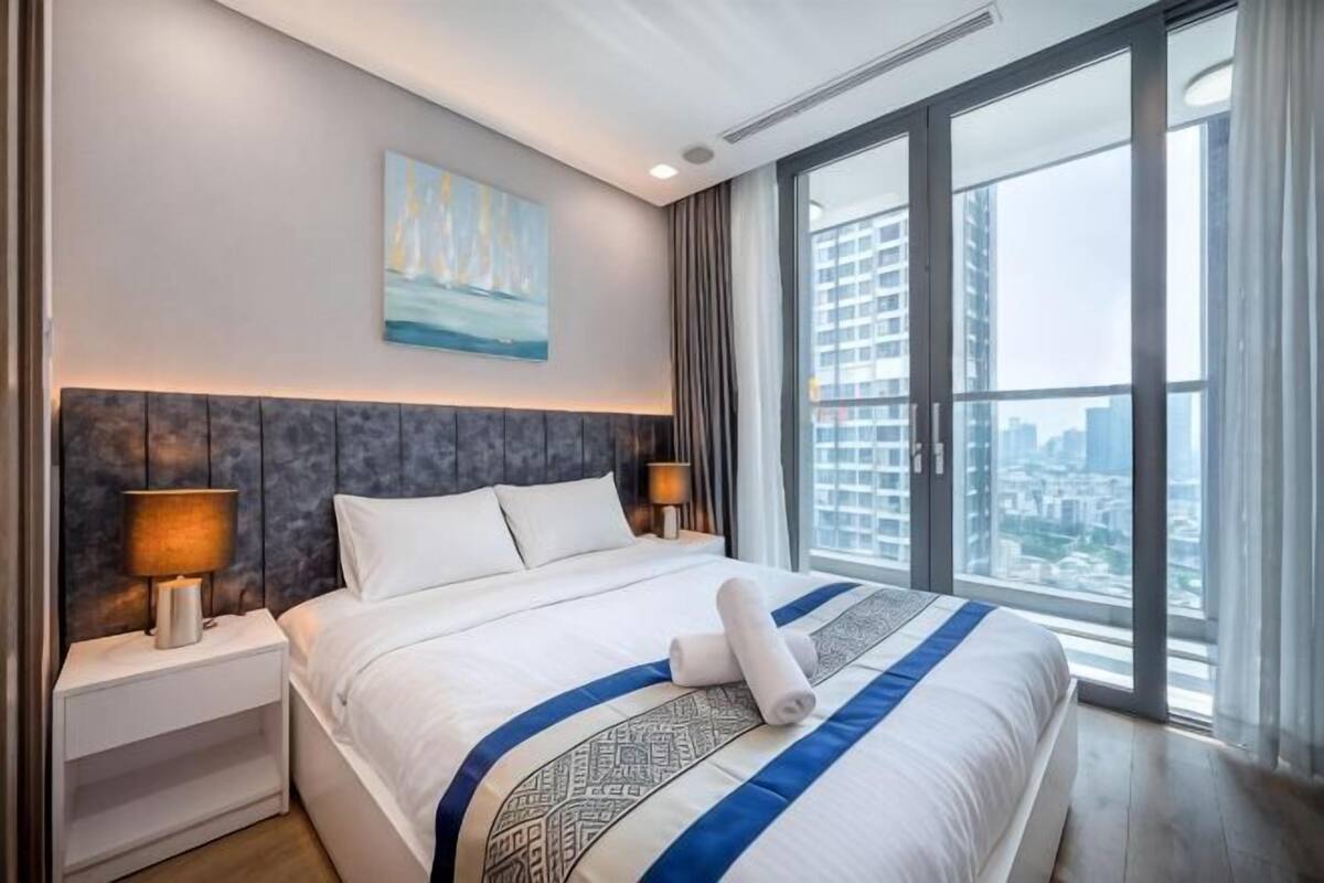A modern hotel room with a large bed, a city view, and a painting on the wall.