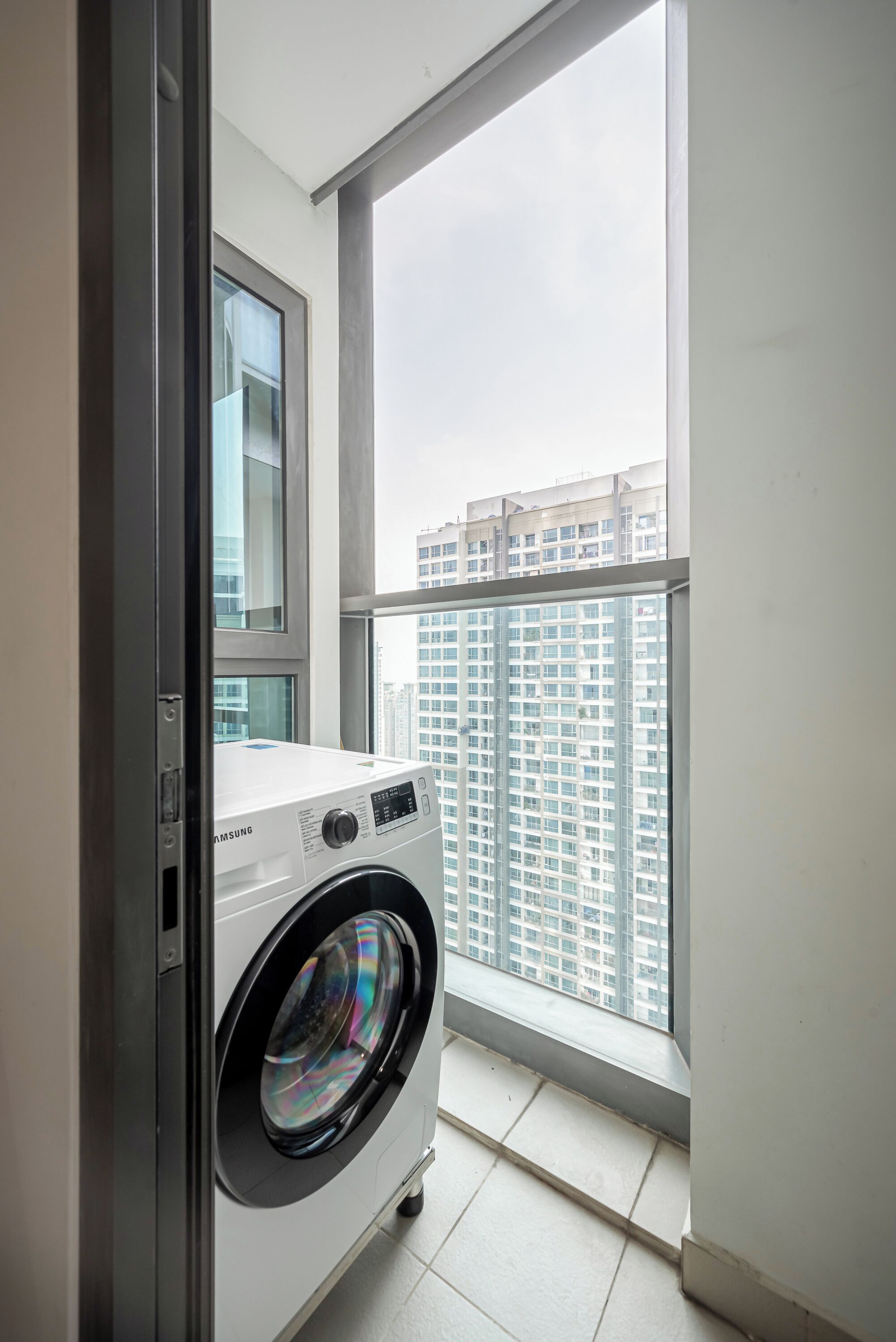 A compact laundry room with a front-loading washing machine, a window, and a view of a high-rise building.