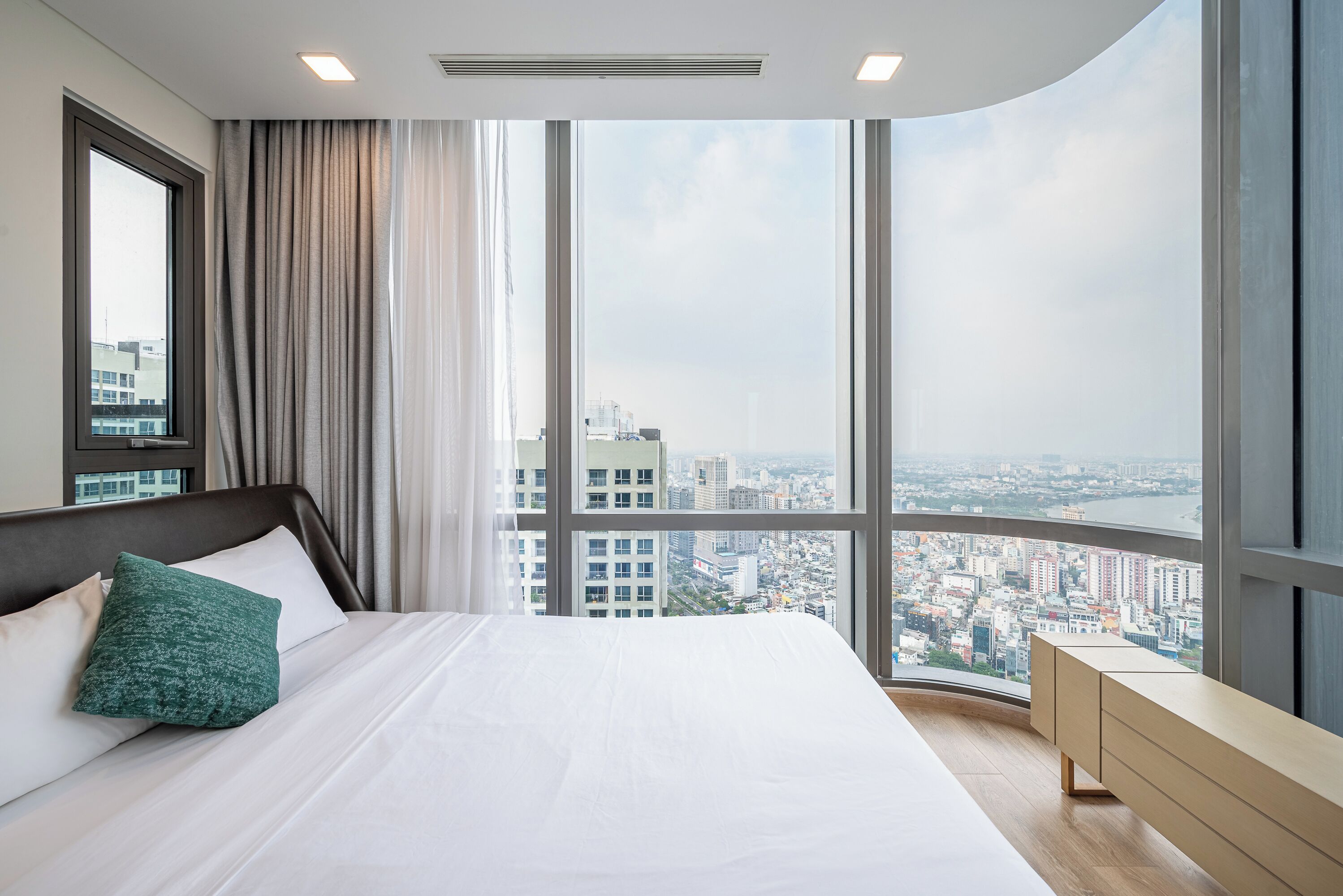 A modern hotel room with a large bed, a nightstand, and a view of a cityscape through large windows.