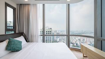 A modern hotel room with a large bed, a nightstand, and a view of a cityscape through large windows.