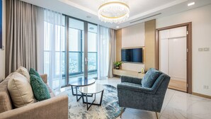 Un salon moderne avec un canapé, un fauteuil et une table ronde. Une grande fenêtre est agrémentée de rideaux, et une suspension orne le plafond. Un téléviseur à écran plat est fixé au mur.