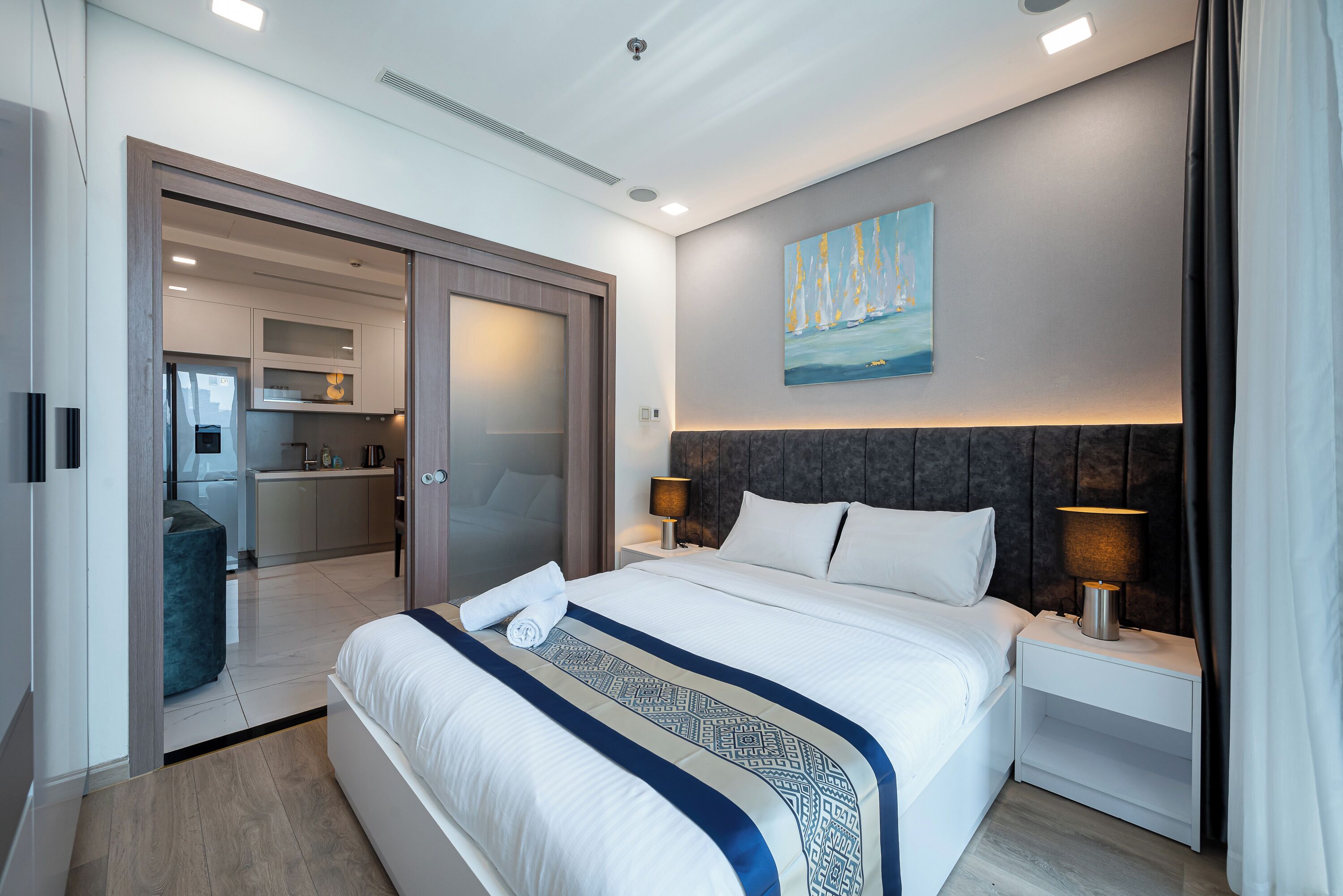 A modern hotel room with a large bed, a bedside table with a lamp, a painting on the wall, and a view into the kitchen area.