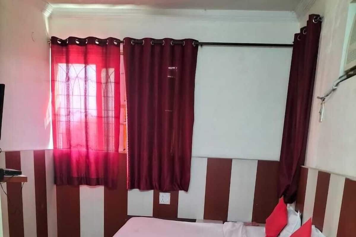 A single-bed room with a wooden headboard, a bedside table with a water bottle, a red cushion, and a window with red curtains.