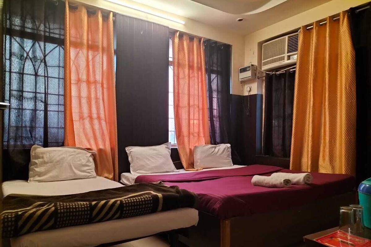 A hotel room with two beds, a window with sheer curtains, an air conditioning unit, and a small table with a red cloth.