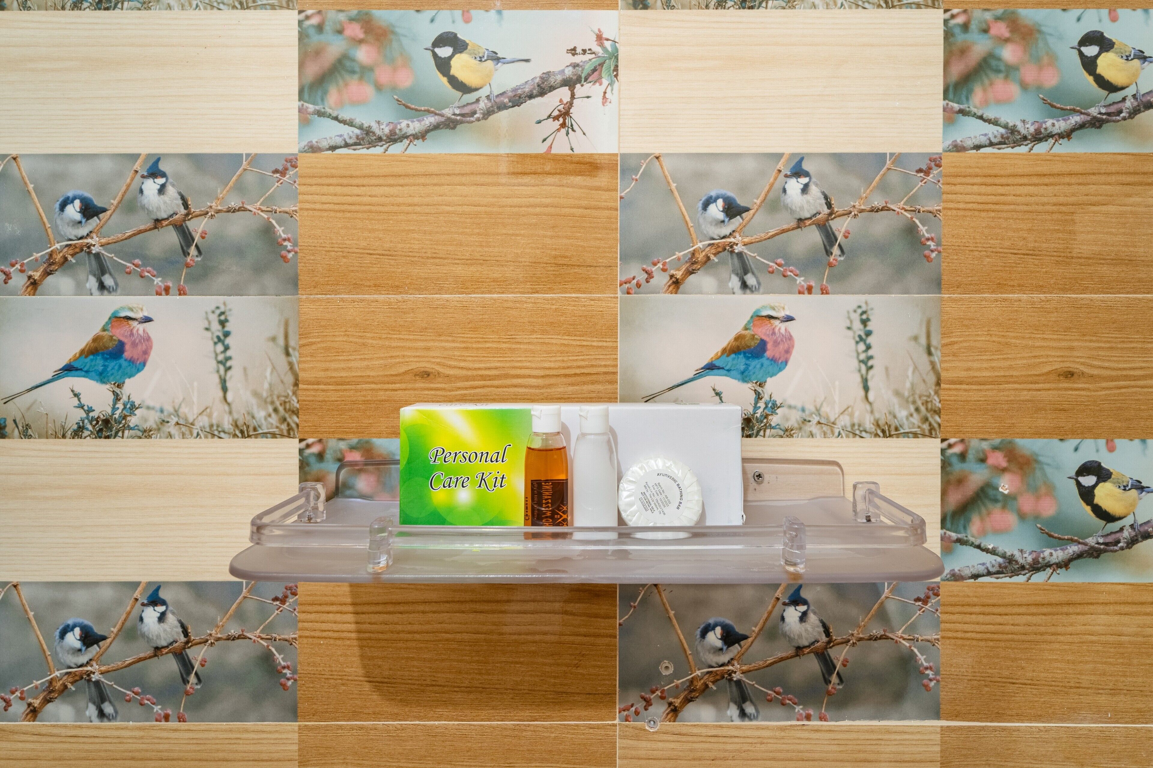 Eine transparente Ablagefläche mit einem Pflegeset, darunter eine grüne Schachtel, Flaschen und ein Behälter, vor einer Wand mit Vogel-Motivfliesen.