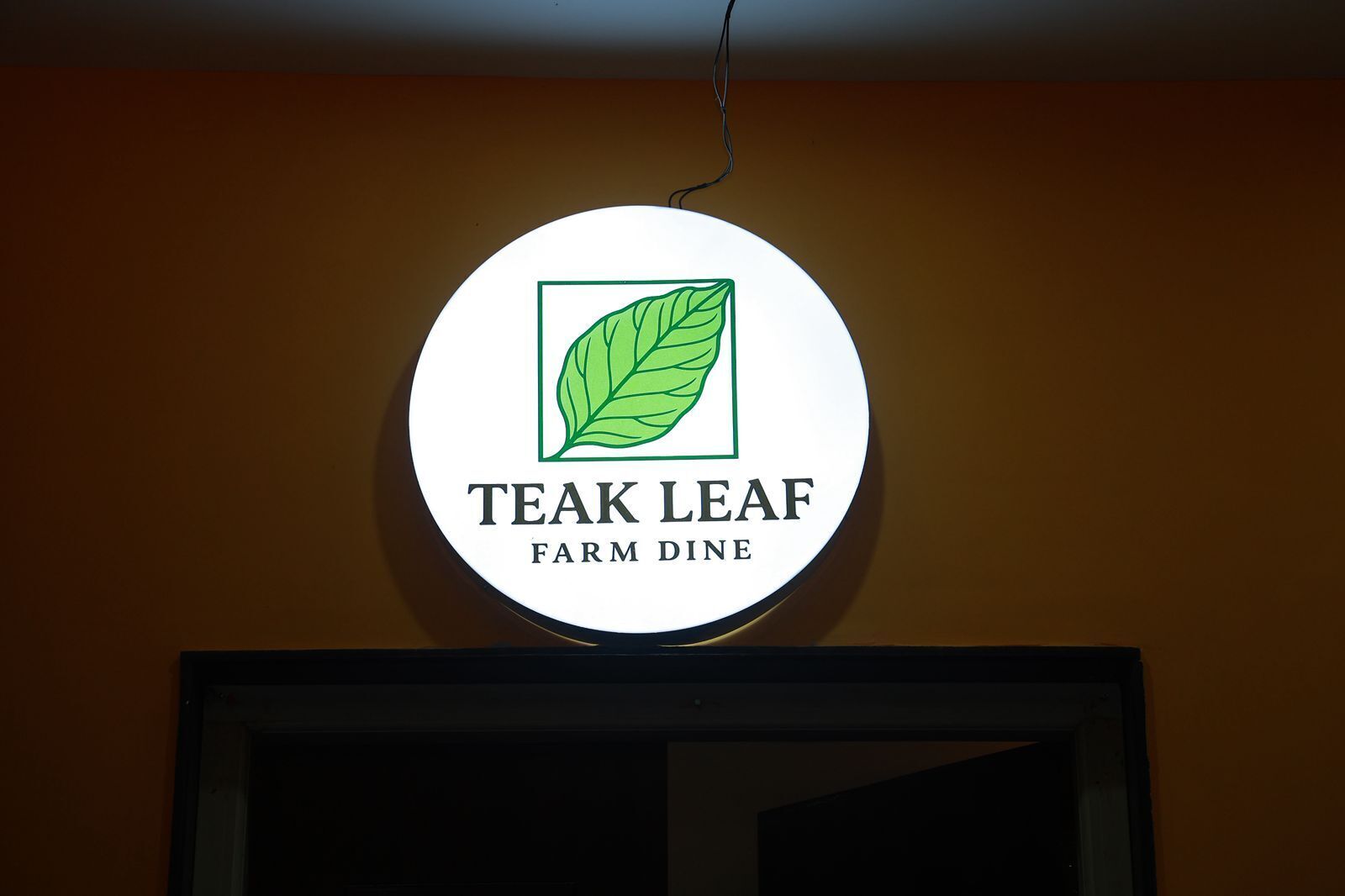 Et rundt skilt med et grønt blad og teksten 'TEAK LEAF FARM DINE' på en væg.