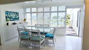 Een eethoek met een glazen tafel en witte stoelen, een groot raam met zonwering en een schilderij aan de muur.