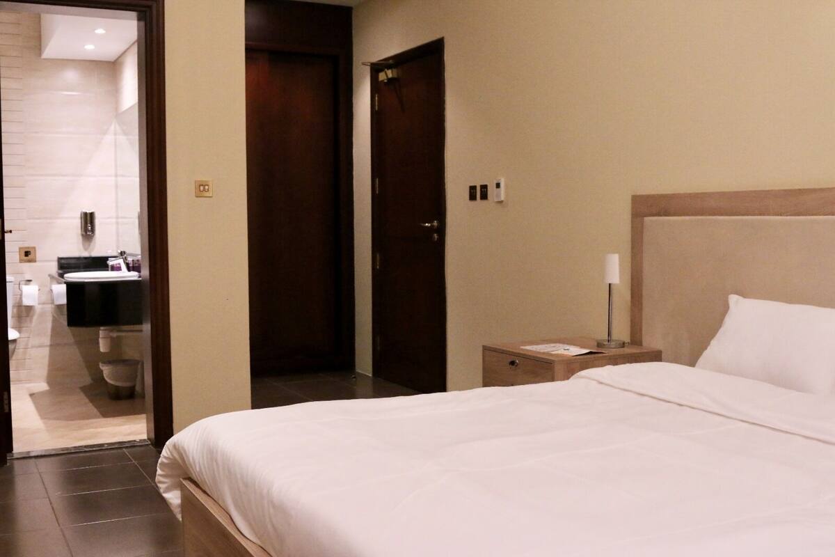 A hotel room with a bed, a nightstand, a lamp, a wooden headboard, and a bathroom visible through an open door.