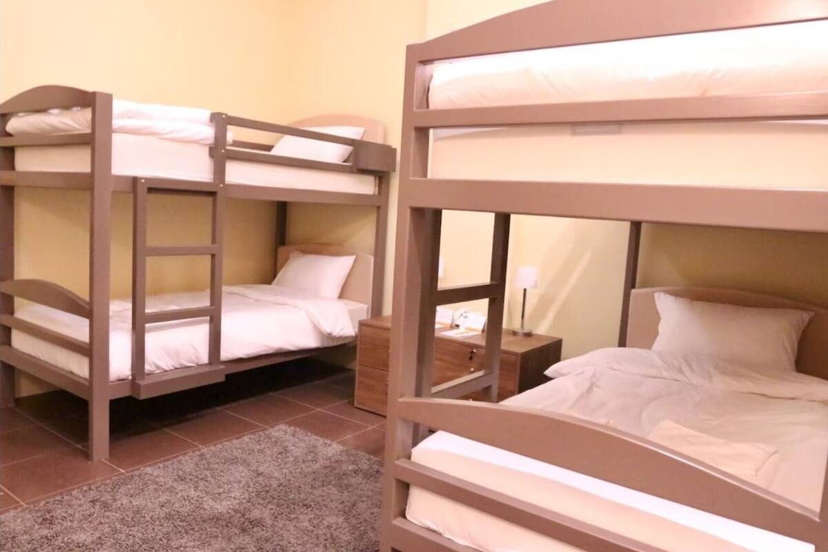 A room with two bunk beds, each with a wooden frame and white bedding. There is a small wooden nightstand with a lamp between the beds.