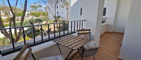 A balcony with a wooden table and two cushioned chairs, a tiled floor, and a view of the surrounding area.