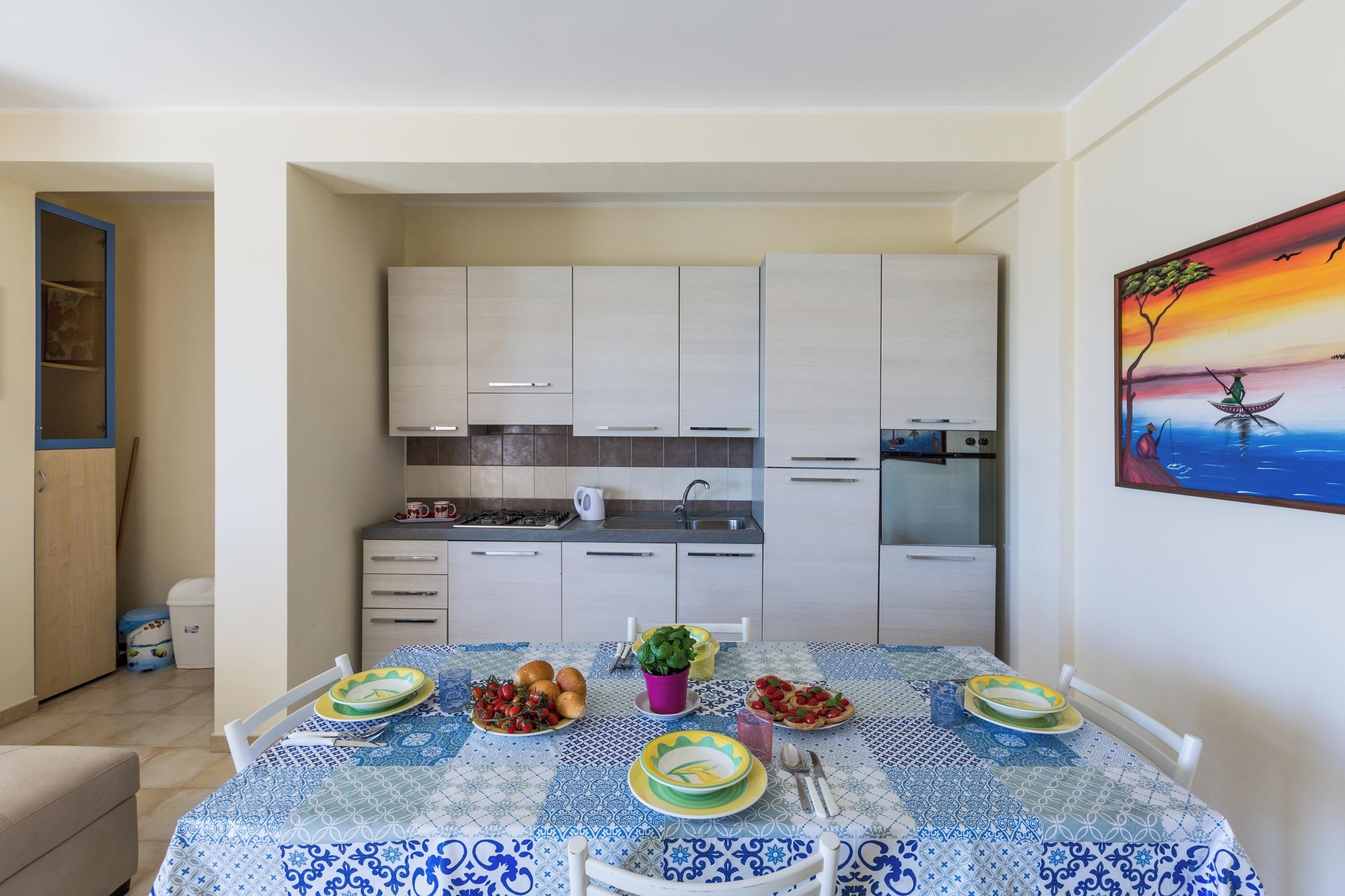 A dining area with a table set for four, a kitchen in the background, and a painting on the wall.
