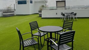 Une terrasse sur le toit avec une pelouse verdoyante, une table ronde entourée de chaises et un bâtiment blanc doté d’une fenêtre et d’une porte.