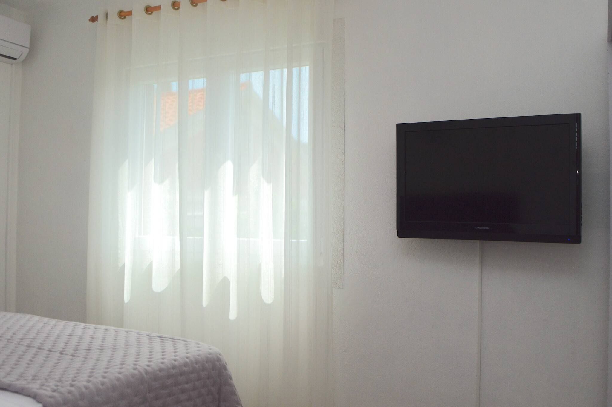 Ein Zimmer mit einem Bett, einem an der Wand befestigten Fernseher und einem Fenster mit leichtem Vorhang.