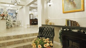 Un hall d’entrée spacieux, agrémenté d’un imposant lustre, d’un fauteuil en cuir noir, d’une table basse en verre sur laquelle est posé un vase de fleurs, d’une cheminée et d’un grand tableau encadré au mur.