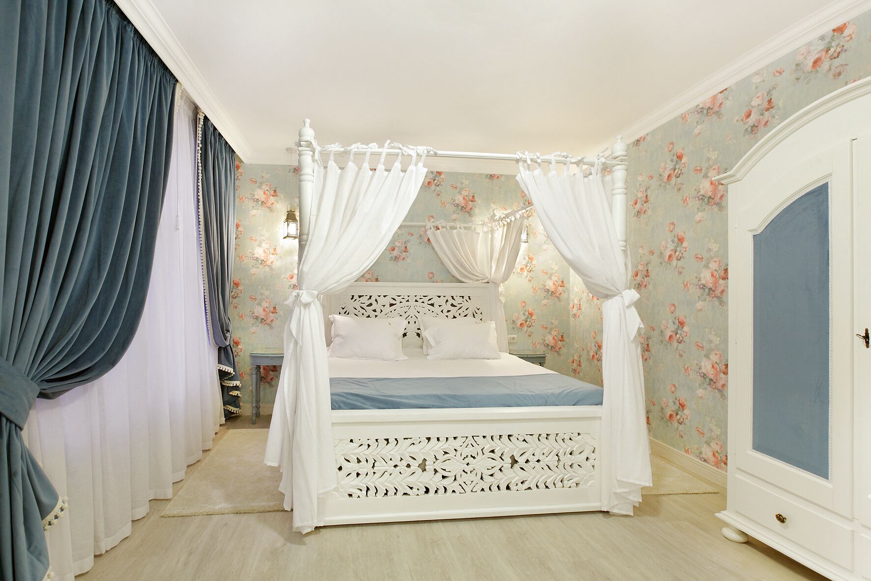 Un lit à baldaquin à quatre poteaux, avec un papier peint à motifs floraux, une armoire blanche et une petite table de chevet.