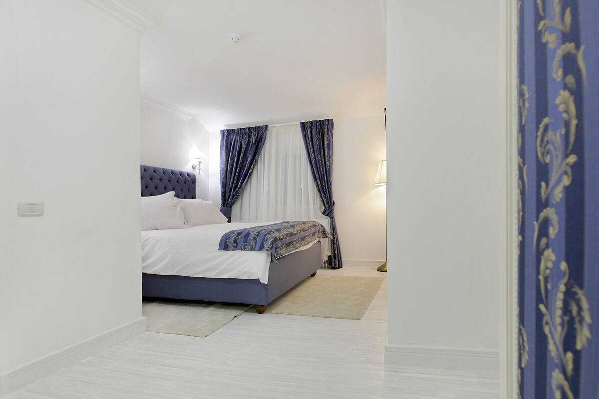 Een slaapkamer met een bed, witte muren, een lichtgekleurde vloer en een gordijn met een blauw-wit patroon.
