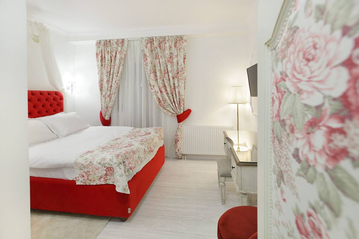 Een slaapkamer met een rood bed, witte gordijnen, een nachtkastje met een lamp en een rode stoel.