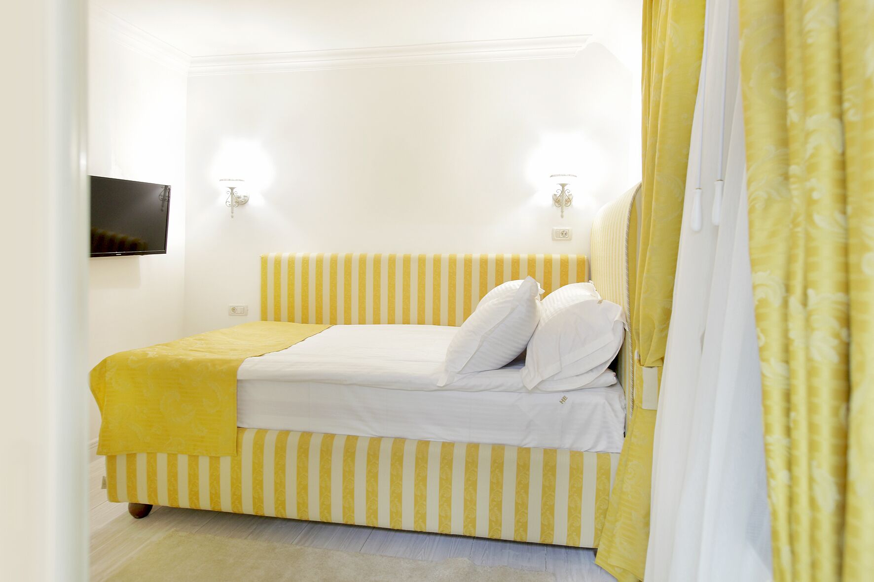 A bed with yellow and white striped bedding, a flat-screen TV on the wall, and a yellow curtain.