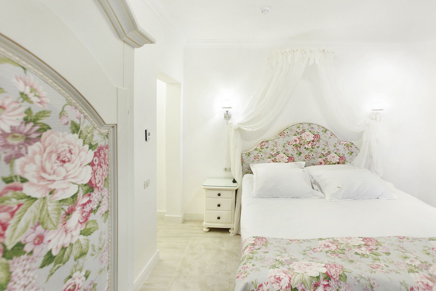 A bedroom with a floral bedspread, a nightstand with drawers, a bedside table, and a canopy bed.