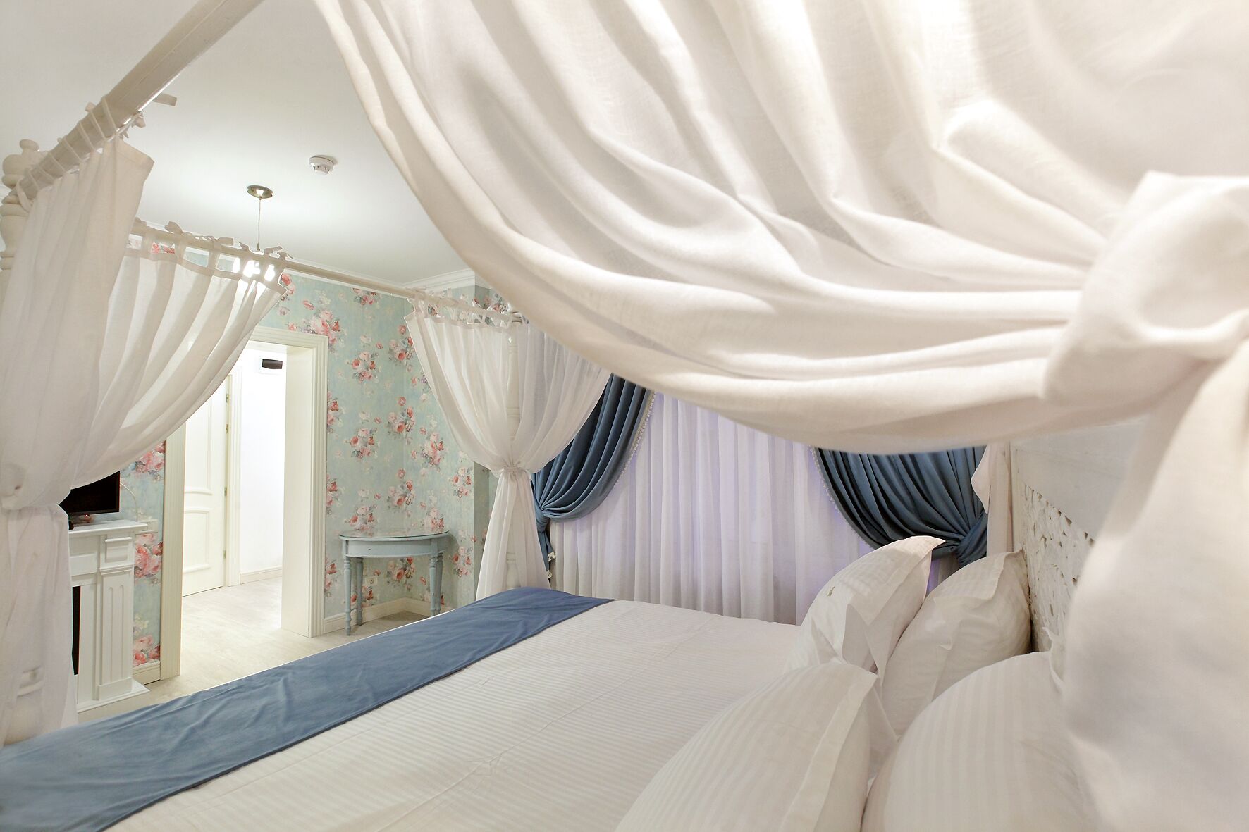 Ein Schlafzimmer mit Baldachinbett, Blumenpapier und einem kleinen Nachttisch, mit Blick auf einen Flur.
