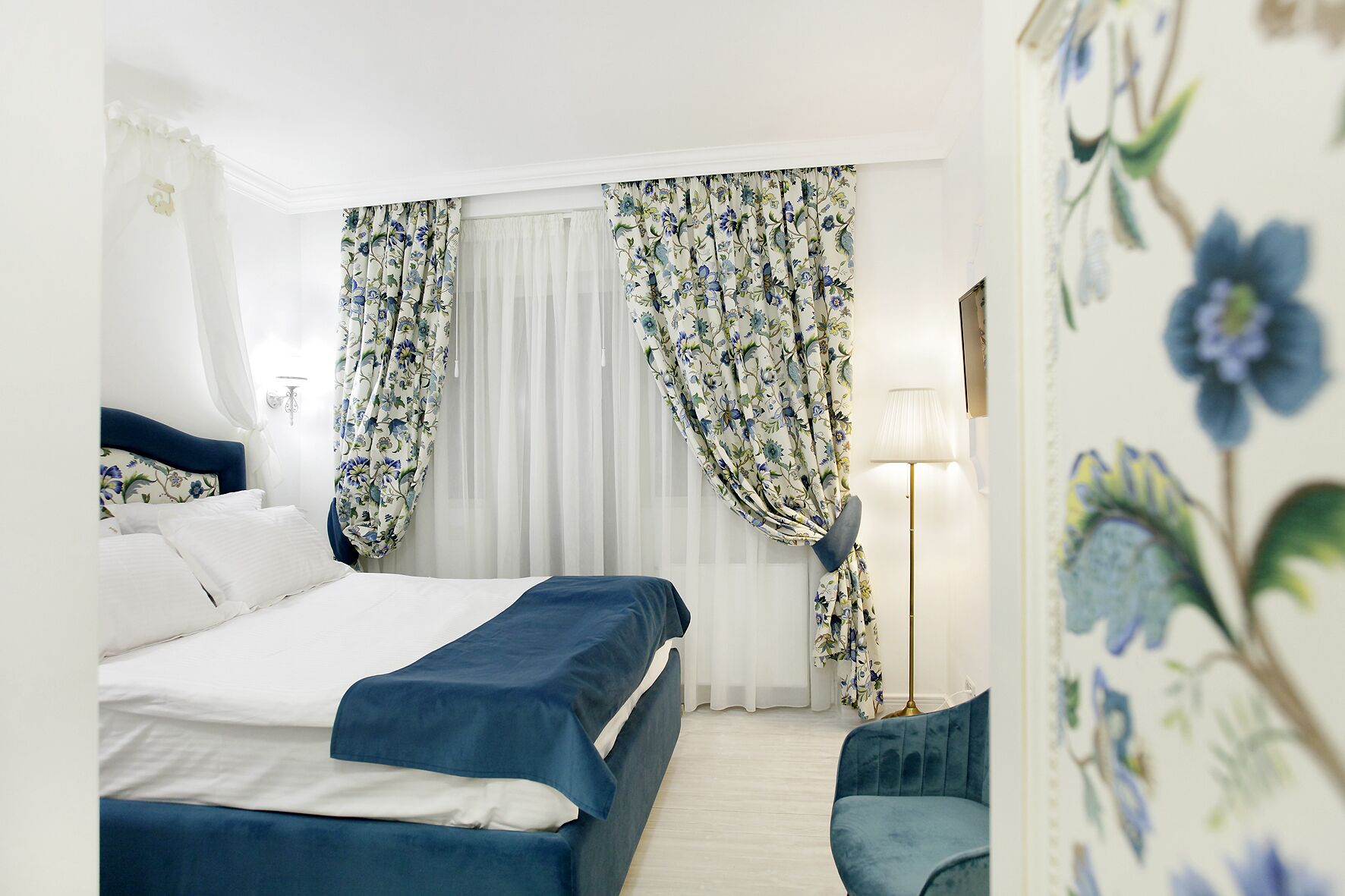 A hotel room with a bed, a chair, a lamp, and floral curtains.