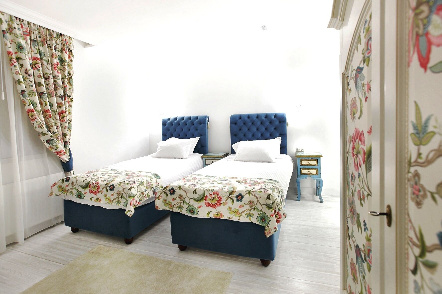 Un dormitorio con dos camas, cortinas florales, un cabecero azul, una mesita de noche pequeña y una puerta con motivos florales.
