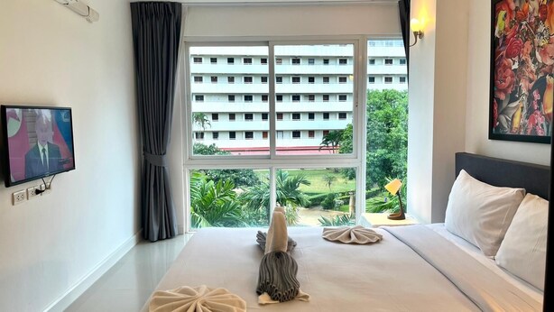 A hotel room with a bed, a TV, a window with a view of a building and greenery, and a painting on the wall.