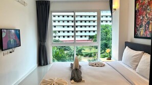 A hotel room with a bed, a TV, a window with a view of a building and greenery, and a painting on the wall. - 6-30 - 1 Bdr Condo with Balcony and shared Pool (Tambon Patong)