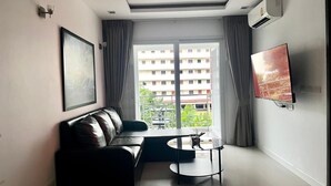 A modern living room with a leather sofa, a small round table, a flat-screen TV mounted on the wall, and a large window with curtains.