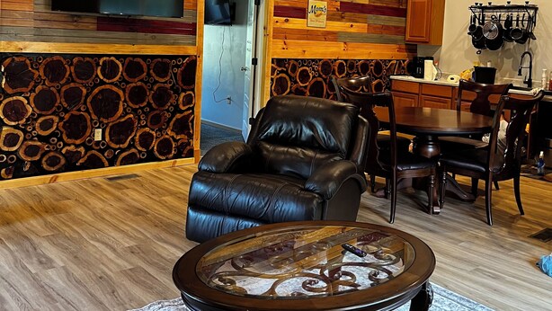 A living room with a fireplace, a leather armchair, a glass coffee table, and a television mounted on a wooden wall.