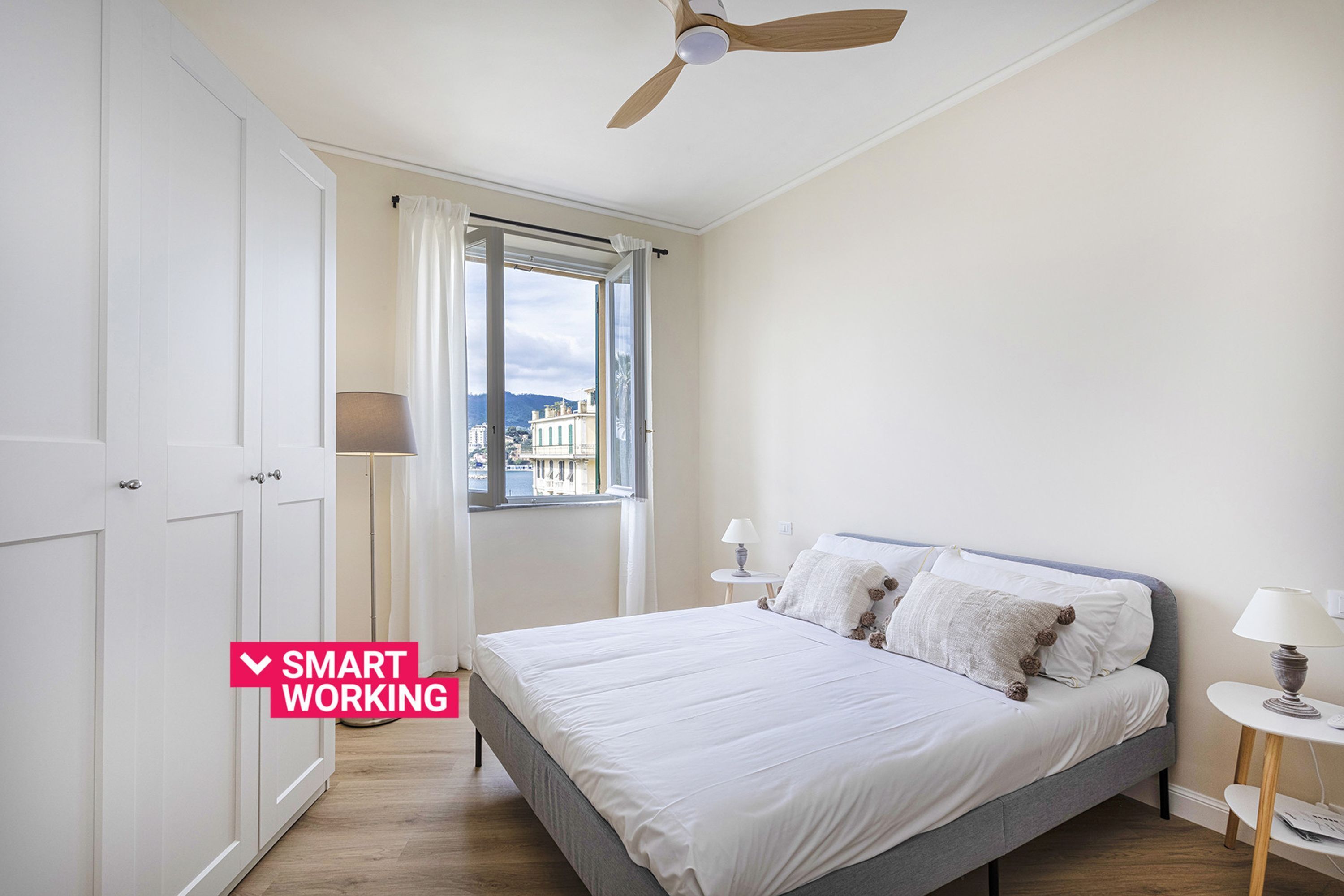 A bedroom with a bed, a ceiling fan, a window with curtains, and a view of buildings outside.