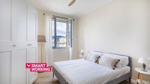 A bedroom with a bed, a ceiling fan, a window with curtains, and a view of buildings outside. - Sea View Flat in Rapallo by Wonderful Italy (Rapallo)