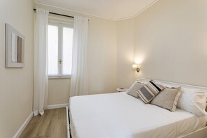 Ein Schlafzimmer mit einem Bett, weißen Vorhängen, einer Nachttischlampe und einem gerahmten Bild an der Wand.
