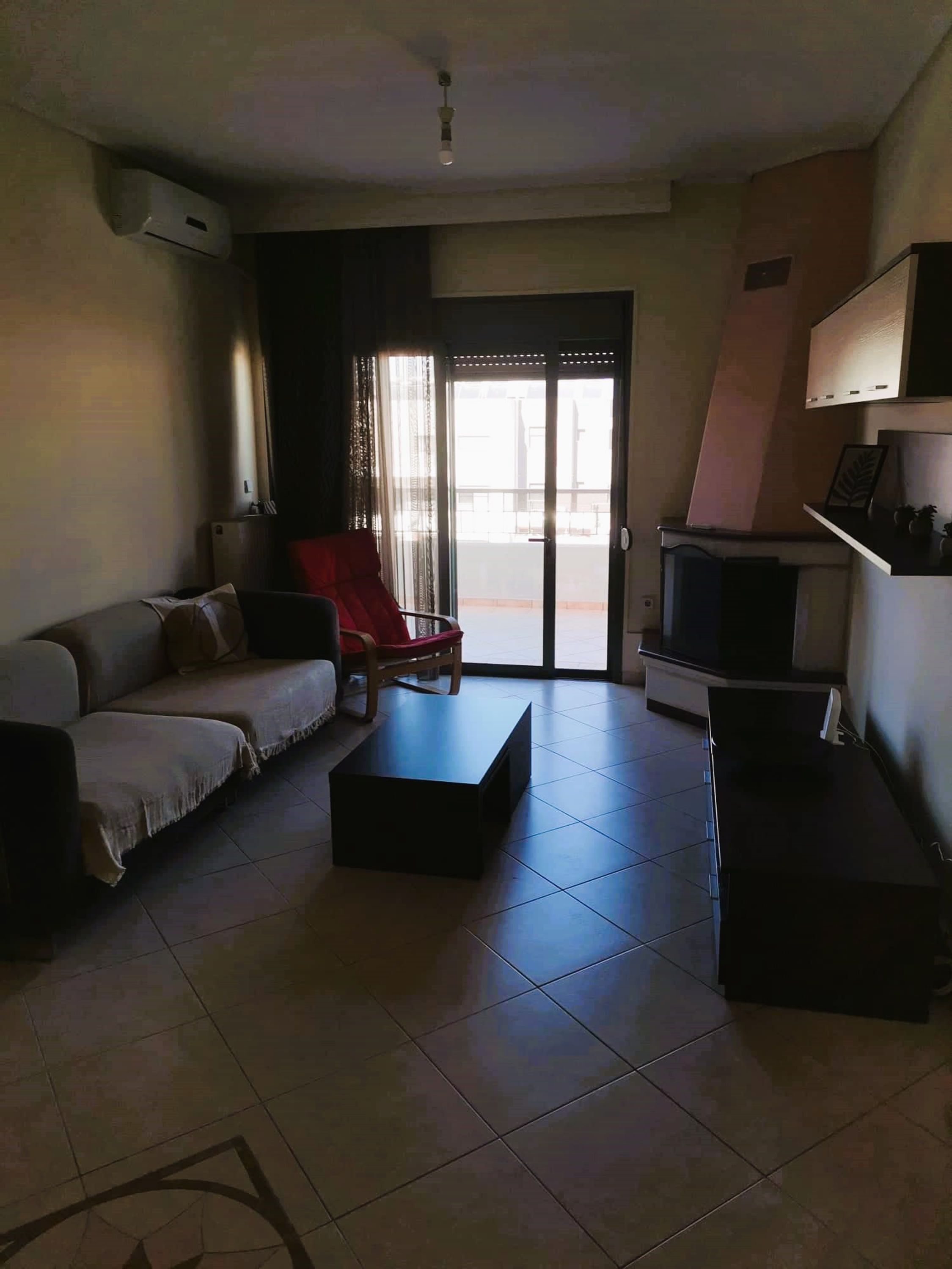 Een woonkamer met een bank, een rode fauteuil, een salontafel, een televisie en een schuifdeur naar een balkon.