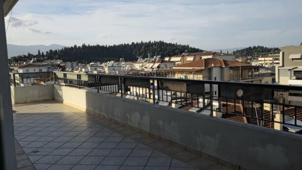 Ein Balkon mit Fliesenboden und Metallgeländer, der auf ein Wohngebiet mit Gebäuden und einen bewaldeten Hügel im Hintergrund blickt.