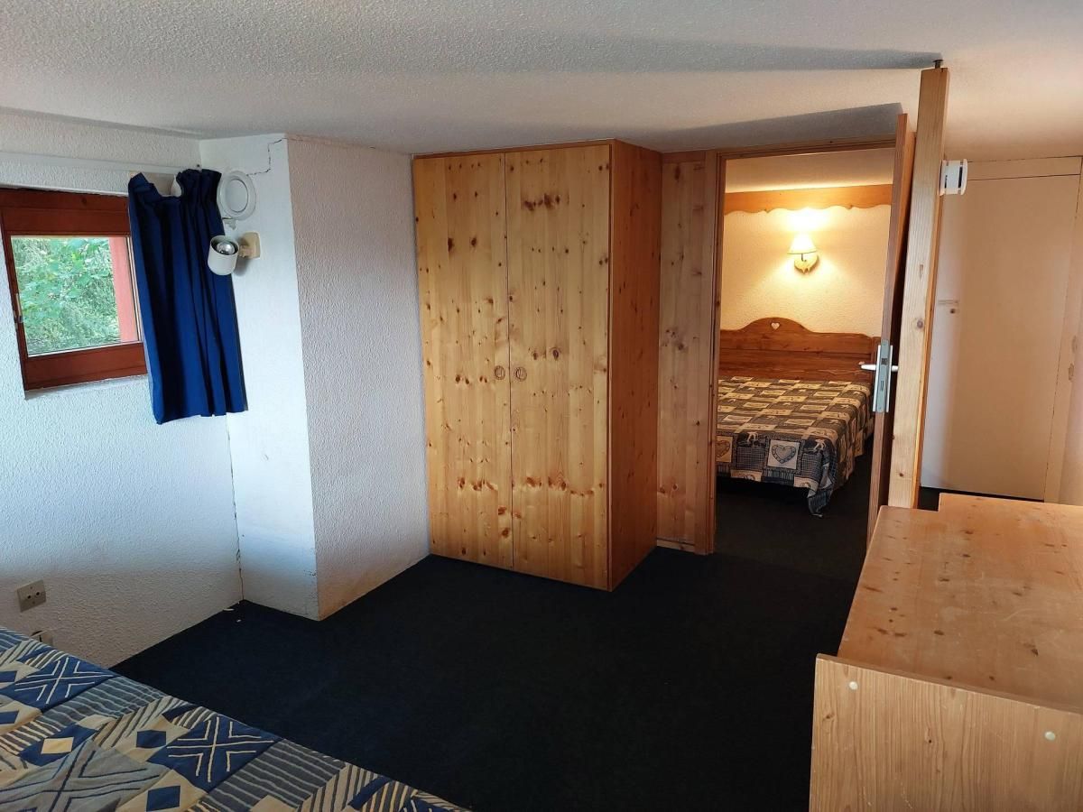 A compact room with a bed, a wooden wardrobe, a small window with curtains, and a door leading to another room.