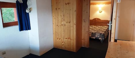 A compact room with a bed, a wooden wardrobe, a small window with curtains, and a door leading to another room.