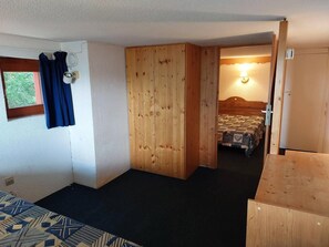 Une pièce de taille modeste comprenant un lit, une armoire en bois, une petite fenêtre avec des rideaux et une porte donnant sur une autre pièce.