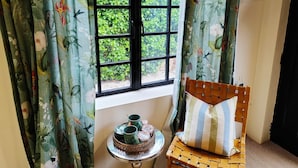 A room with a window, curtains, a small table with a tray of items, and a chair with a cushion. - Cotswold Bankview (Moreton-in-Marsh)