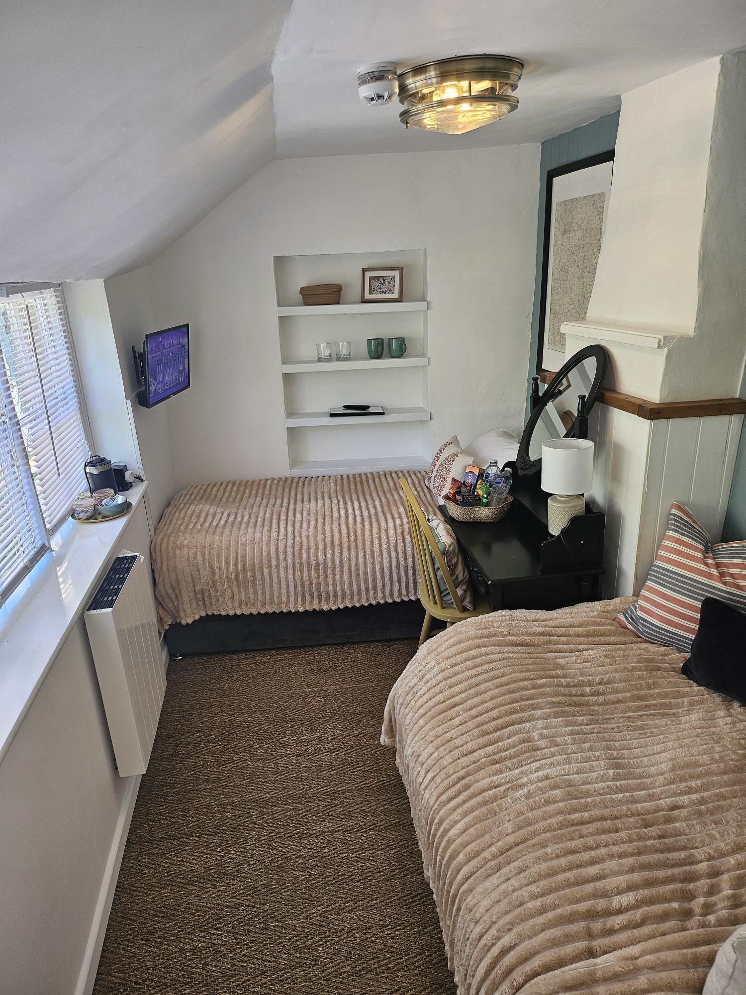 A hotel room with two beds, a TV, a shelf with items, and a window with blinds.