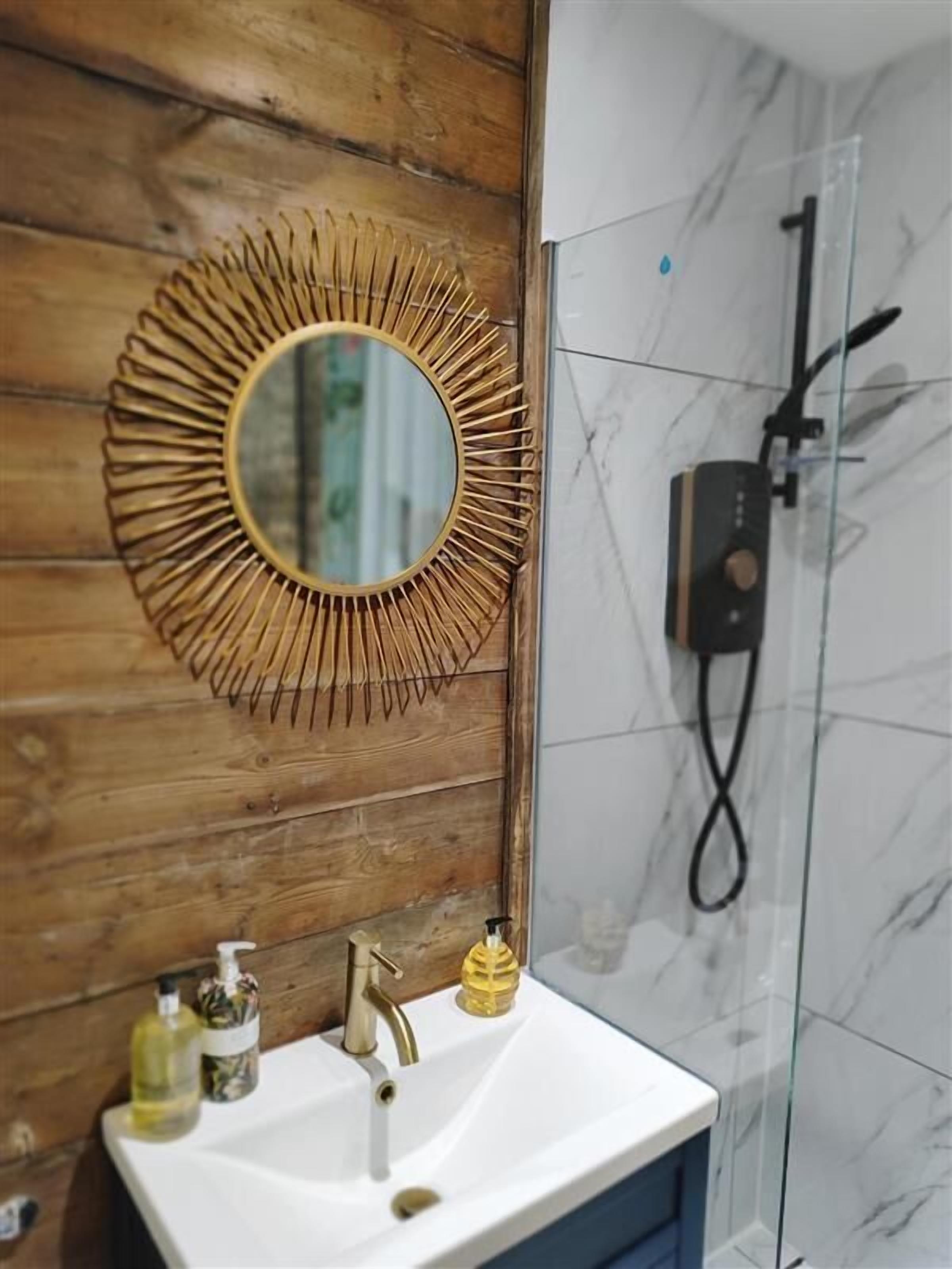 A bathroom with a wooden wall, a round mirror with a sunburst frame, a white sink, and a shower with a glass door.