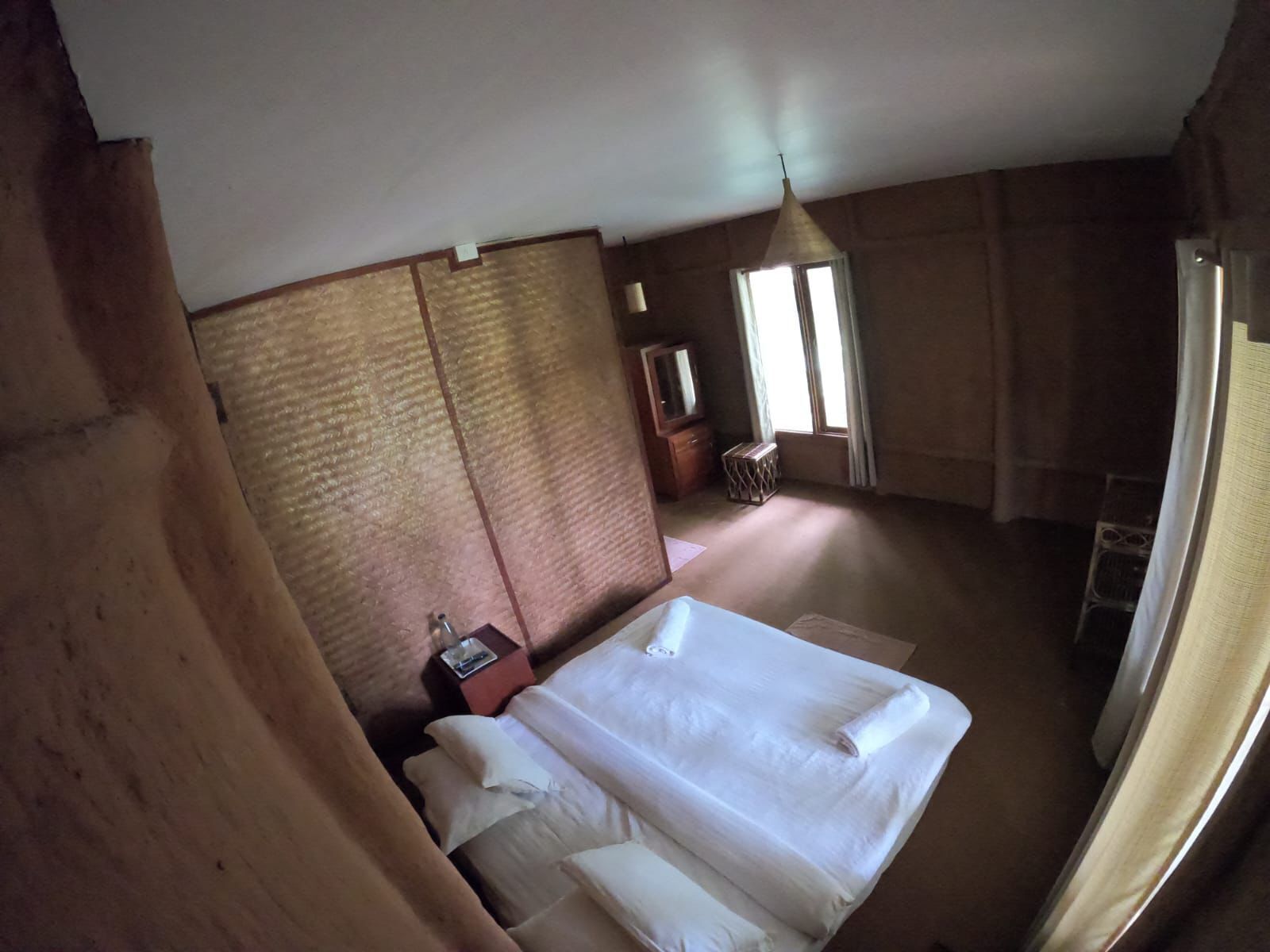 Una habitación de hotel con una cama grande, una mesita de noche, una pequeña mesa, una ventana con cortinas y una lámpara colgante.