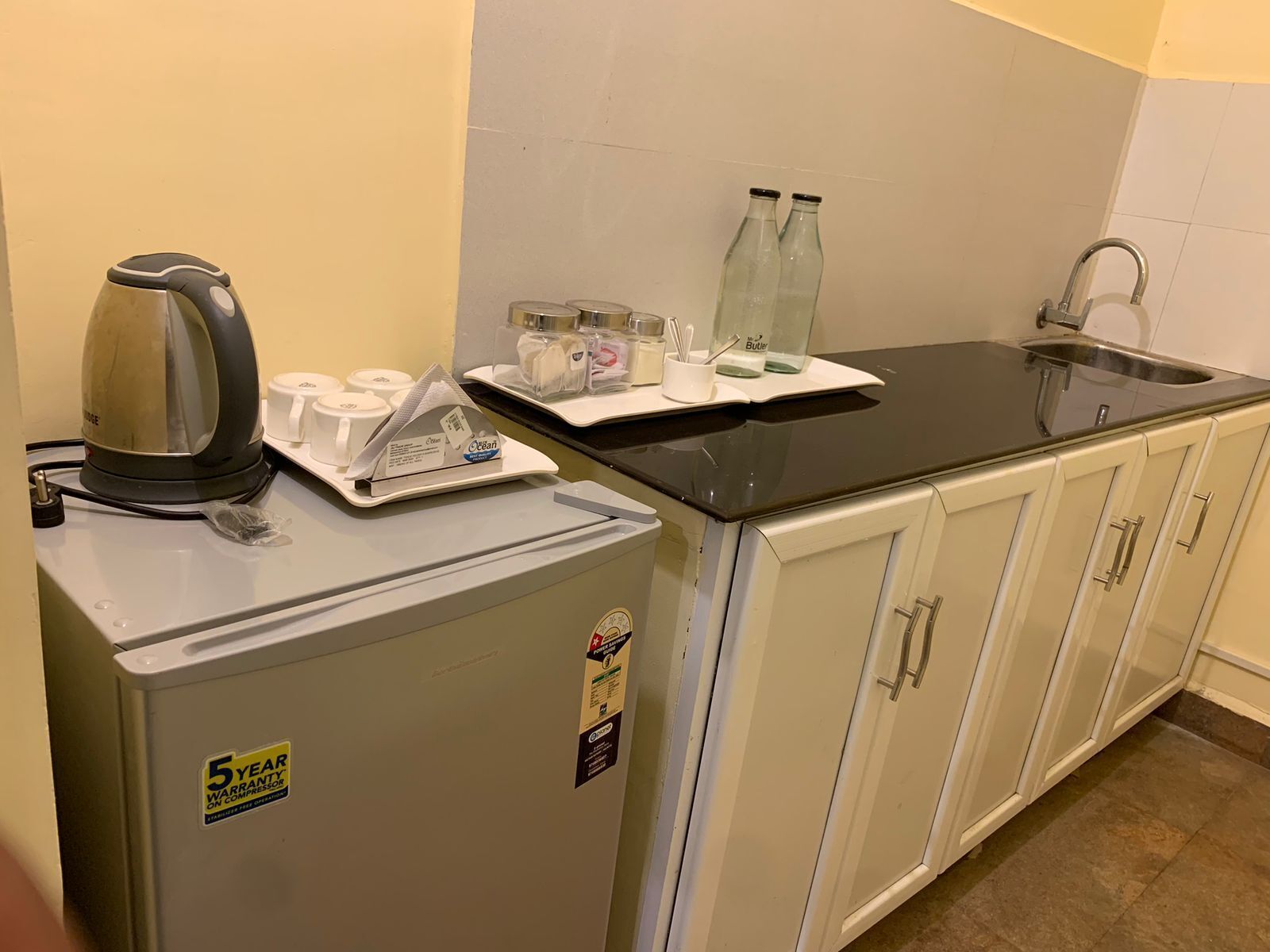 Una kitchenette compacta con cafetera, un refrigerador pequeño, un lavamanos y una encimera con varios objetos.