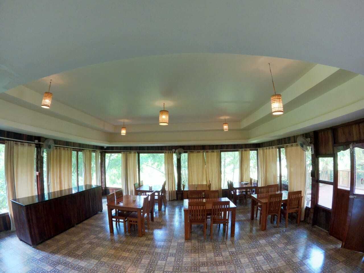 A spacious dining area with multiple wooden tables and chairs, large windows with curtains, and pendant lighting.