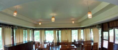 A spacious dining area with multiple wooden tables and chairs, large windows with curtains, and pendant lighting.
