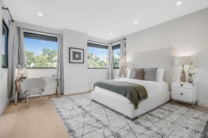 A modern bedroom with a large bed, bedside tables, a desk, and a view of greenery outside.