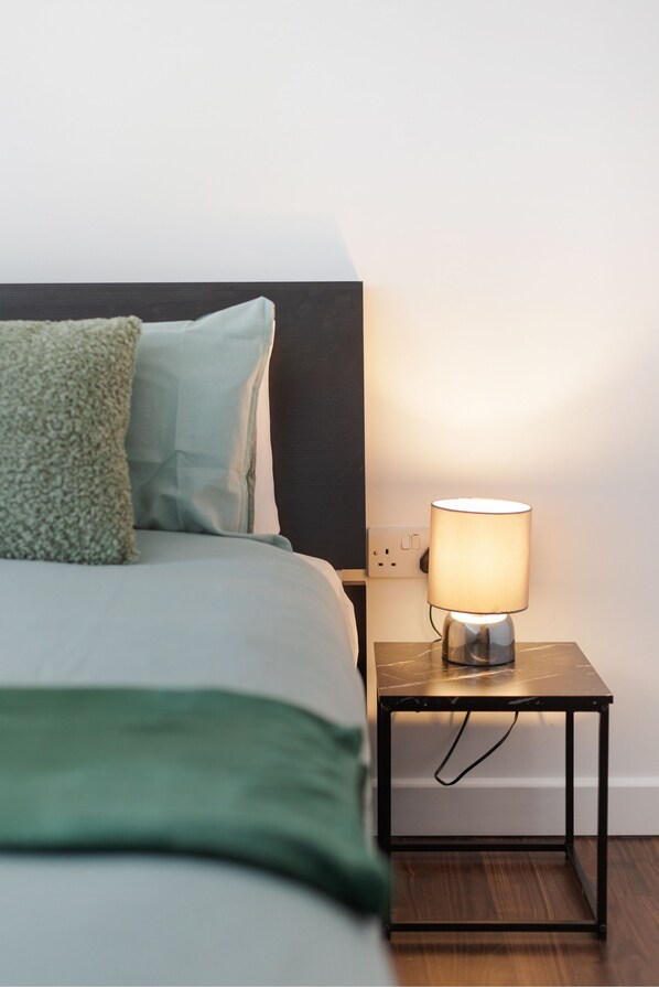 A neatly made bed with a green throw blanket and pillows, a bedside lamp on a small table, and a power outlet on the wall.